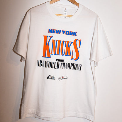 A white short-sleeve t-shirt hanging on a wooden hanger against a plain light wall. The shirt features a large front graphic celebrating the New York Knicks as 2025 NBA World Champions, with 'NEW YORK' in blue, 'Knicks' in orange and blue, and '2025 NBA WORLD CHAMPIONS' in black.