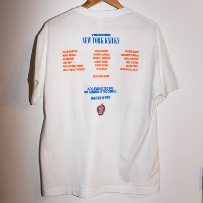 A white short-sleeve t-shirt hanging on a wooden hanger against a plain light wall. The back of the shirt features a large printed graphic with the text 'NEW YORK KNICKS' in blue, followed by two columns of red and blue names, and a small New York Knicks logo near the bottom.