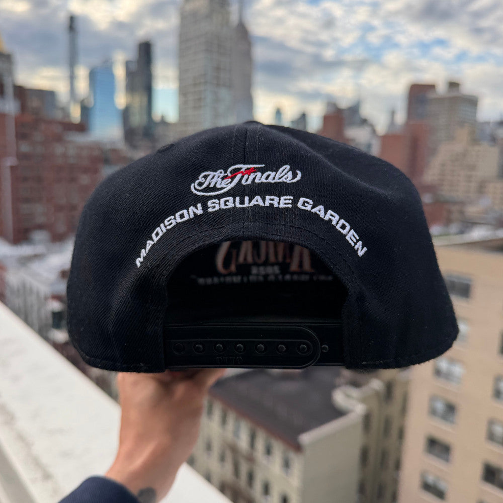 A black baseball cap held up by a hand, featuring white embroidered text reading 'The Finals' and 'MADISON SQUARE GARDEN' on the front, with a blurred city skyline in the background.