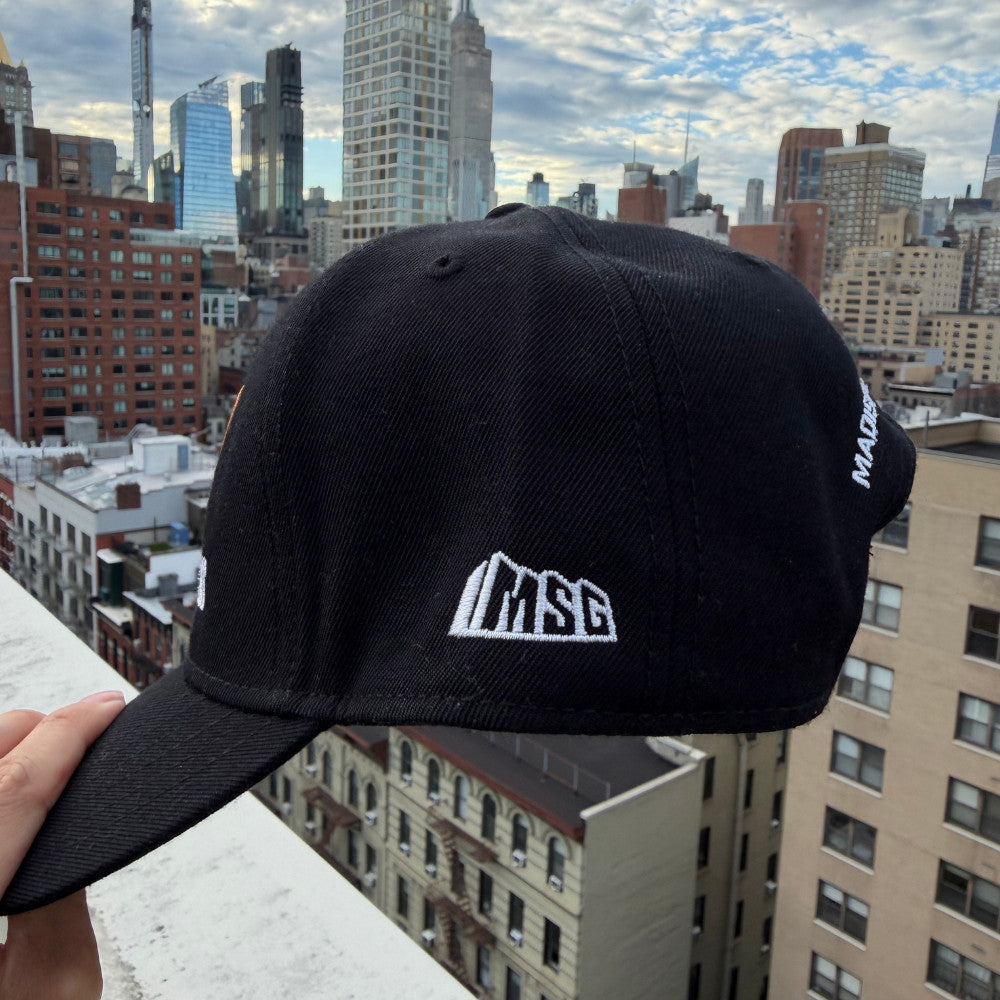 A black baseball cap with a white embroidered 'MSG' logo on the front, held up by a hand against a city skyline background.