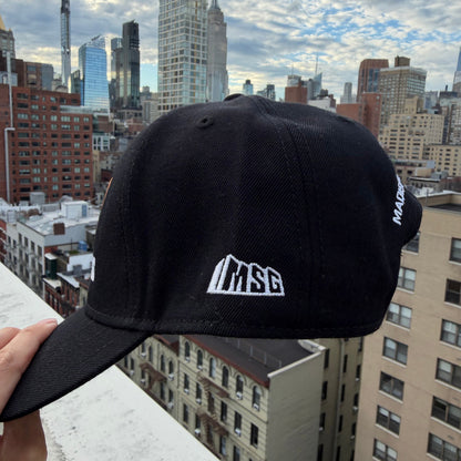 A black baseball cap with a white embroidered 'MSG' logo on the front, held up by a hand against a city skyline background.
