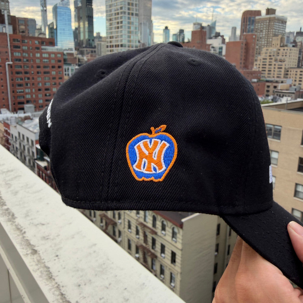 A black baseball cap held up by a hand, featuring an embroidered New York Yankees logo inside an apple outline on the front, with a city skyline in the background.